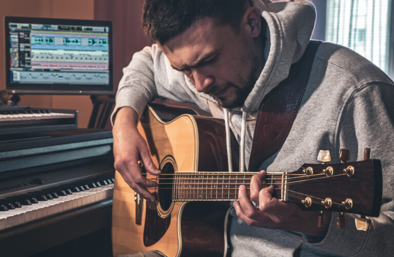 Male Musician Plays The Guitar At Home In The Workplace Near The Computer.
